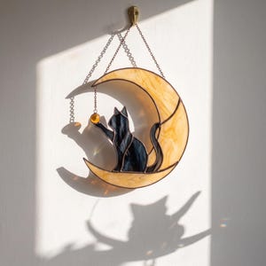 May include: A stained glass suncatcher featuring a black cat sitting on a crescent moon made of marbled cream and yellow glass. The cat reaches for a small, round, amber-colored glass bead suspended by a metal chain. The piece hangs from a metal chain.