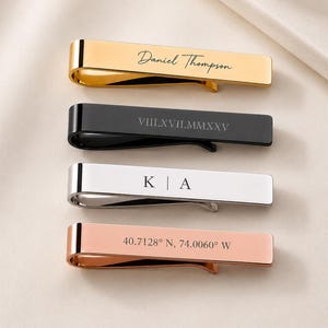 May include: Four metal tie clips in gold, black, silver, and rose gold tones. The gold clip is engraved with the name Daniel Thompson, the black clip with VIII.XVII.MMXXV, the silver clip with K | A, and the rose gold clip with 40.7128° N, 74.0060° W.