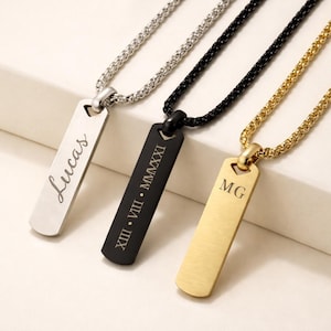 Peut inclure: Trois colliers pendentifs personnalisés. L'un est argenté avec le nom "Lucas" gravé, un autre est noir avec des chiffres romains, et le troisième est doré avec les initiales "MG". Chaque collier a une chaîne.