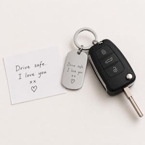 Könnte beinhalten: Ein silberfarbener Metall-Schlüsselanhänger mit einer Gravur des Textes Drive safe.. I love you xx und einem Herzsymbol, befestigt an einem Metallring mit einem schwarzen Autoschlüssel. Ein weißer quadratischer Zettel zeigt die gleiche handschriftliche Nachricht.