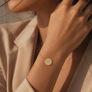 May include: A delicate gold bracelet with a circular charm engraved with the letter "B". The bracelet is displayed on a wrist, against a soft, neutral background. The jewellery is simple and elegant.