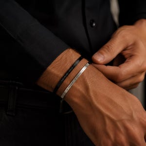 Könnte beinhalten: Zwei dünne Metallarmreifen, einer in Mattschwarz und einer in Silber, jeweils mit Textgravur. Der schwarze Armreif trägt die Worte DISCIPLINE | FOCUS | FREEDOM, während der silberne Armreif mit den Worten BE PRESENT | BE STRONG | BE YOU graviert ist.