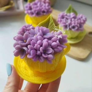 May include: A yellow teacup-shaped soap with a purple flower arrangement. The soap has a saucer and a green leaf. The text "Surprise Savon" is visible. The soap is held by a person. The background is blurred.