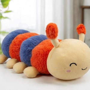 May include: A plush caterpillar toy featuring a smooth cream-coloured head with a smiling face, two orange fuzzy antennae, and a segmented body alternating between textured blue and orange sections with small cream-coloured feet along the base.