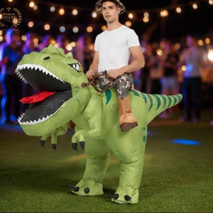 May include: An inflatable green Tyrannosaurus Rex costume featuring a wide-open mouth with white teeth and a red tongue. The costume includes a brown saddle and reins, with attached faux legs wearing brown boots, creating the appearance of riding.