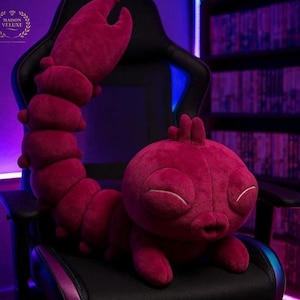 May include: A plush, magenta-colored scorpion toy with a segmented body, large claw, and a curled tail. The scorpion has closed eyes and is resting on a black chair. The image includes the text "MAISON VELUXE".
