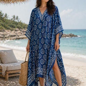 Può includere: Un abito caftano blu e bianco a fantasia con scollo a V e spacco laterale. L'abito è realizzato in tessuto leggero ed è indossato da una persona che porta una borsa tote intrecciata. Lo sfondo comprende una spiaggia, l'oceano e un ombrellone di paglia.