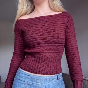 Könnte beinhalten: Ein dunkelroter Langarmpullover mit weitem schulterfreiem Ausschnitt und einer texturierten horizontalen Häkelmusterstruktur. Das Kleidungsstück verfügt über einen eng anliegenden gerippten Taillenbereich und lange Ärmel, kombiniert mit einer hellblauen Jeans.