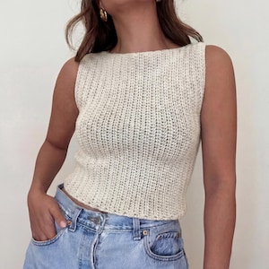 May include: Cream-colored, sleeveless, knitted top with a ribbed texture. The top has a boat neck and a fitted silhouette. The model is wearing light blue denim jeans. The top appears to be handmade, showcasing a classic, timeless design.