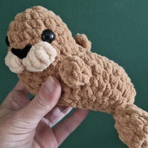Puede incluir: Un pequeño peluche de foca tejido a ganchillo en color marrón claro con un acabado de hilo suave y texturizado. La foca presenta dos ojos redondos de plástico negro, una pequeña nariz negra bordada y un área de hocico en color beige claro.