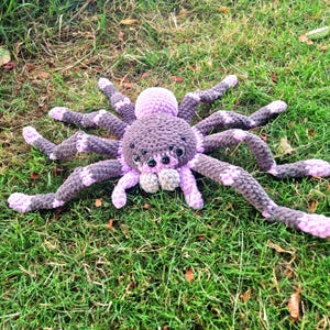 Pode incluir: Um brinquedo de pelúcia de aranha feito de crochê com um corpo redondo e oito pernas longas. O fio é uma mistura de cinza escuro e roxo claro, com detalhes em roxo claro nas pontas das pernas e articulações. Possui vários olhos pretos.