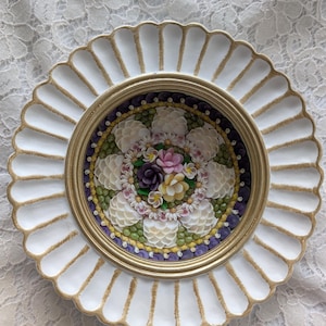 May include: A white ceramic plate with a gold rim and a scalloped edge. The center of the plate features a mosaic of seashells arranged in a floral design with pink, purple, and white flowers.