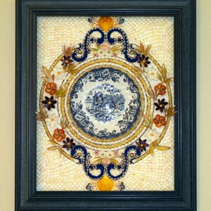 May include: Framed artwork featuring a blue and white porcelain plate with a pastoral scene, surrounded by a decorative arrangement of shells, flowers, and embellishments. The artwork is encased in a dark blue frame.
