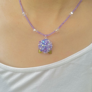 May include: A necklace featuring a lavender beaded chain with a pendant made of purple and blue beads. The pendant is adorned with small green leaf details. Clear beads are spaced along the chain.
