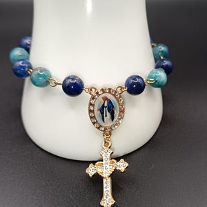 Op de afbeelding: Een rozenkransarmband met blauwe en blauwgroene kralen. De armband heeft een goudkleurig middelpunt met een afbeelding en een gouden kruisvormige hanger met heldere accenten. De kralen zijn geregen op een goudkleurige ketting.