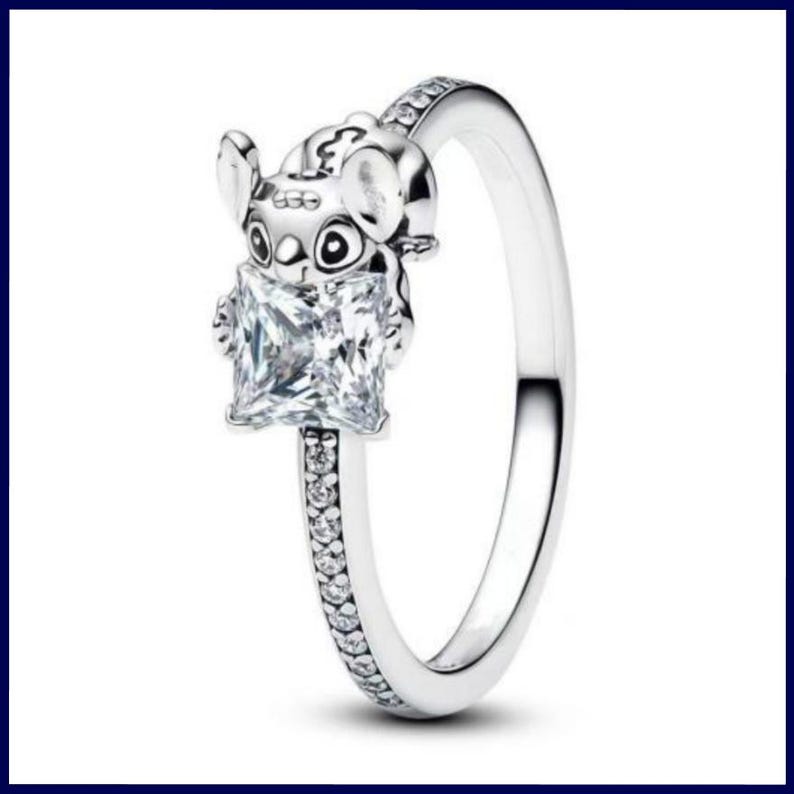 May include: A silver ring featuring a cartoon character holding a square, clear gemstone. The ring band is embellished with small, clear stones. The character has large eyes and ears. The ring is set against a white background.