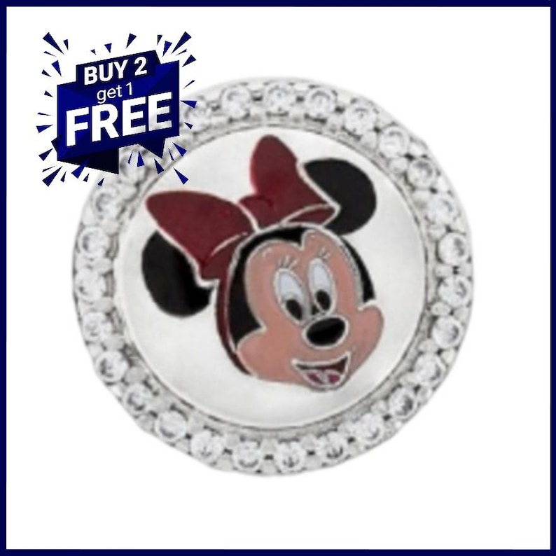 K&ouml;nnte beinhalten: Ein runder, silberfarbener Charm-Anh&auml;nger mit einem Minnie Mouse-Design, das eine rote Schleife und ein rosa Gesicht zeigt. Der Charm ist von einem Ring aus klaren Kristallen umgeben. Der Text "BUY 2 get 1 FREE" ist ebenfalls enthalten.