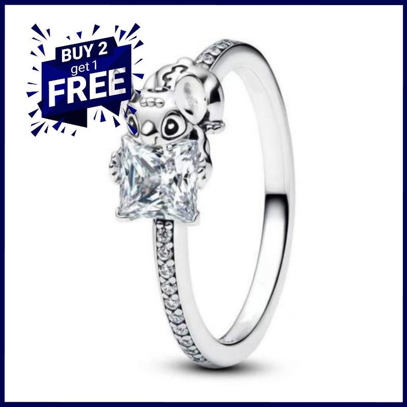 May include: A silver ring featuring a cartoon character holding a large, square, clear gemstone. The ring band is embellished with small, sparkling stones. The image includes a blue and white graphic with the text "BUY 2 get 1 FREE."