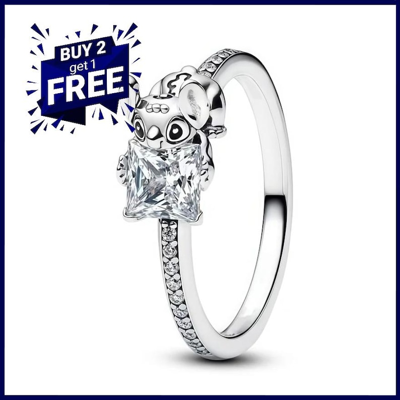 Puede incluir: Un anillo plateado con un personaje de Stitch sosteniendo una gema cuadrada y transparente. La banda del anillo est&aacute; adornada con peque&ntilde;as piedras transparentes. Un gr&aacute;fico azul y blanco en la esquina superior izquierda dice "BUY 2 get 1 FREE".