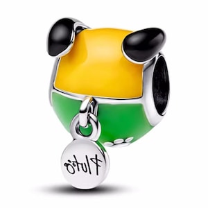 Könnte beinhalten: Ein Charm mit einem hundeähnlichen Gesichtsdesign. Der Charm hat einen gelben Kopf, einen grünen Körper und schwarze Ohren. Ein kleines, rundes Etikett mit Text hängt an dem Charm. Der Charm ist aus silberfarbenem Metall.