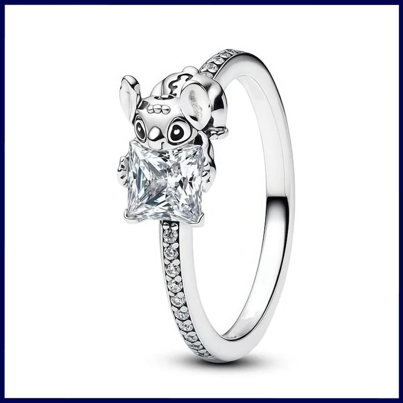 Puede incluir: Un anillo plateado con un peque&ntilde;o personaje de Stitch sosteniendo una gema cuadrada y transparente. La banda del anillo est&aacute; adornada con peque&ntilde;as gemas transparentes. El anillo est&aacute; sobre un fondo blanco.