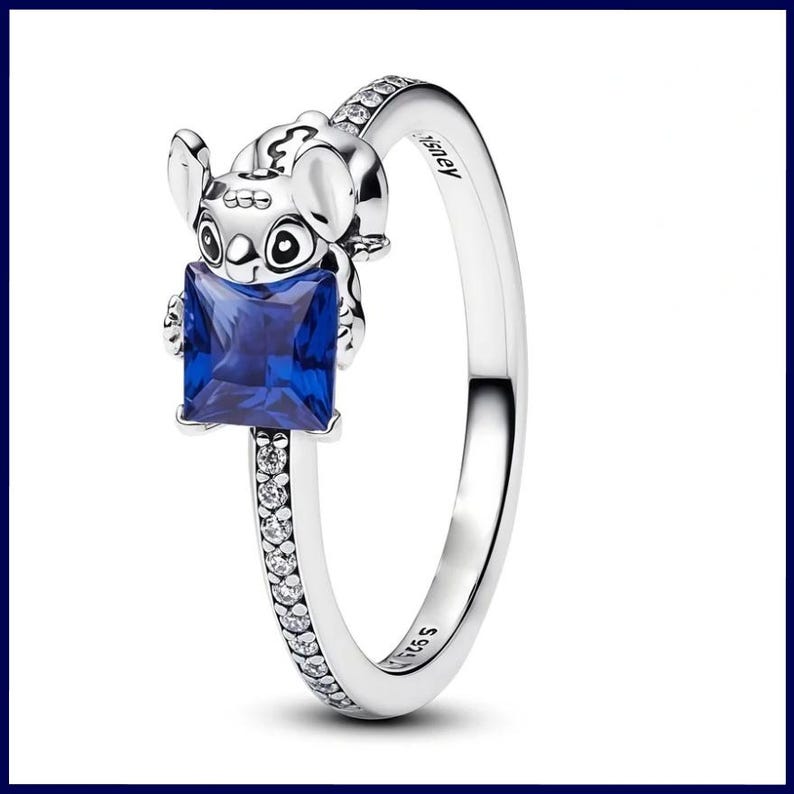 May include: A silver ring with a Stitch figure holding a square, deep blue gemstone. The ring band is decorated with small, clear stones. The word "Disney" is engraved on the ring. This is a piece of jewellery.