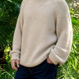 Peut inclure: Un pull beige à manches longues avec un col rond épais et côtelé ainsi que des poignets assortis. Le vêtement présente une texture en tricot épais et une coupe décontractée, porté avec un jean bleu foncé. Le pull est d'une couleur unie.