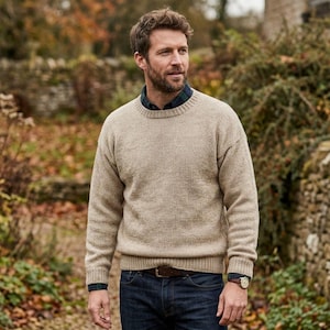 May include: A beige crew neck sweater with long sleeves. The sweater is worn with a plaid shirt underneath, blue denim jeans, and a brown leather belt. A wristwatch is visible.