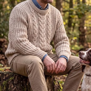 Könnte beinhalten: Ein beigefarbener Zopfmuster-Pullover mit geripptem Rundhalsausschnitt und Bündchen, getragen über einem blauen Hemd mit Kragen, kombiniert mit einer beigen Hose. Der Pullover hat ein strukturiertes vertikales Muster mit Zopfstrick.