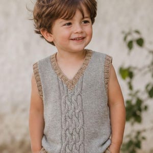 May include: A sleeveless knit sweater vest in light grey featuring a central vertical cable knit pattern. The garment has a V-neckline and ribbed trim in a contrasting tan colour along the neckline, armholes, and bottom hem, creating a textured look.