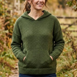 May include: A green hooded sweater with a kangaroo pocket. The sweater is made of a knitted material and is worn with blue jeans. The background is a blurred outdoor scene with trees and a wooden fence.