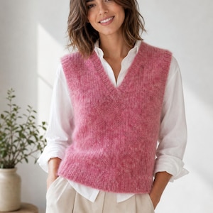 Puede incluir: Un chaleco sin mangas con cuello en V confeccionado en un tejido de punto rosa texturizado y difuso. La prenda presenta un cuello, sisas y dobladillo acanalados, superpuesto sobre una camisa blanca de manga larga y pantalón beige claro.