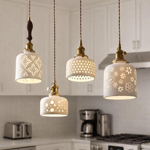 Puede incluir: Cuatro lámparas colgantes con pantallas de cerámica blanca y accesorios de latón cuelgan en una cocina. Las pantallas presentan varios patrones recortados, incluidos diseños florales y geométricos. Los cables son trenzados marrones.