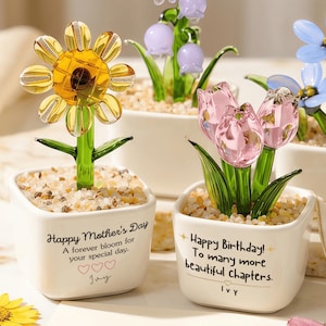 May include: Four decorative glass flower arrangements in white ceramic pots. The flowers include a yellow sunflower, purple bell-shaped flowers, pink tulips, and blue flowers. The pots have text that reads "Happy Mother's Day" and "Happy Birthday!"