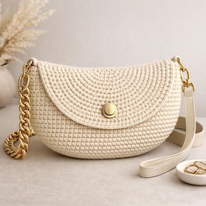 以下が含まれることがあります： クリーム色のかぎ針編みクロスボディバッグ。ゴールドのチェーンストラップと、お揃いのゴールドの留め金が付いています。バッグは丸みを帯びた形で、フラップ開閉です。小さなオフホワイトのレザーストラップも付属しています。スタイリッシュなアクセサリーです。