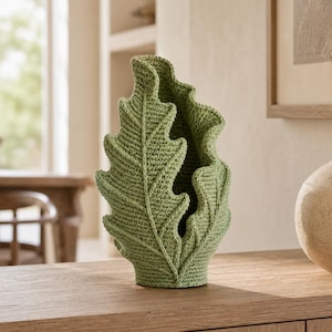 May include: A sage green crochet vase shaped like a large, upright oak leaf. The object features a dense, textured stitch pattern with a prominent central vein and ruffled, wavy edges that create a hollow, open center for holding items.