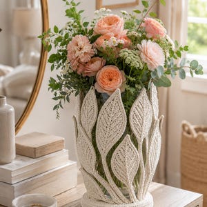 May include: A clear glass vase encased in a cream-colored, crocheted sleeve featuring a pattern of upright, textured leaves. The vase is filled with a bouquet of peach-colored garden roses, small white flowers, and green eucalyptus foliage.