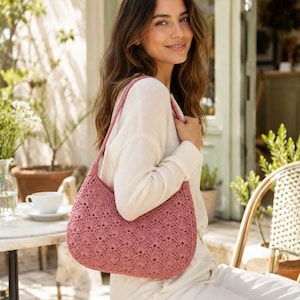 Pode incluir: Uma bolsa de ombro de crochê em rosa antigo com um padrão de pontos repetitivos em forma de leque. A bolsa apresenta uma silhueta curva com uma alça única integrada, feita de fio texturizado em um tom rosa suave e sólido em todo o design.