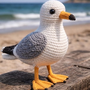 Könnte beinhalten: Handgehäkelte Möwe mit weißem Körper, grauen Flügeln und gelben Füßen. Der Schnabel ist schwarz und gelb. Sie steht auf einer Holzoberfläche, mit Strand und Meer im Hintergrund.