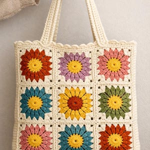 May include: Cream-colored crochet tote bag featuring nine colorful flower squares. Each square has a yellow center and petals in shades of orange, purple, pink, blue, and green. The bag has a scalloped edge and sturdy handles.