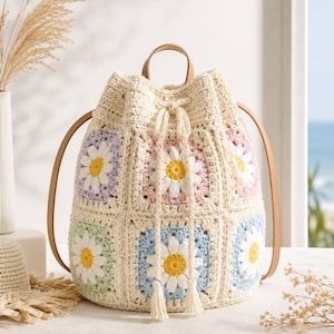 Può includere: Una borsa a secchiello all'uncinetto con base color crema e pannelli quadrati che presentano motivi a margherita in tonalità pastello di lavanda, rosa, verde chiaro e blu. La borsa include cinturini in similpelle beige e chiusura a coulisse con nappe.