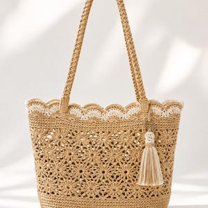 Può includere: Una borsa tote all'uncinetto beige con un motivo floreale e manici intrecciati. La borsa presenta un bordo smerlato con finiture bianche e un ciondolo a nappa. La borsa è progettata per trasportare oggetti.