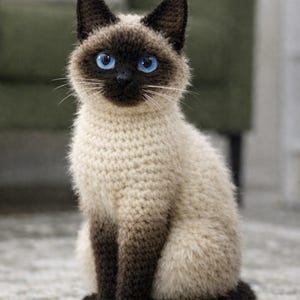 以下が含まれることがあります： 鮮やかな青い目を 가진 かぎ針編みのシャム猫の置物。猫はクリーム色の体に、耳、足、尾に濃い茶色のアクセントがあります。猫は模様のあるラグの上に座っています。
