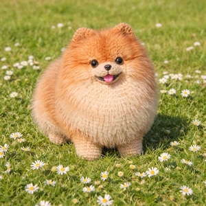 May include: A crocheted Pomeranian dog with a fluffy, orange-tan coat and a smiling expression. The dog is positioned on a bed of green grass dotted with small white daisies. The dog's tongue is pink.