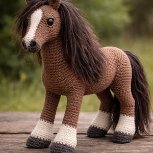 May include: A crocheted brown horse with a white stripe down its face and a long, dark mane and tail. The horse has white and gray hooves and is posed on a wooden surface. The background is a blurred green.