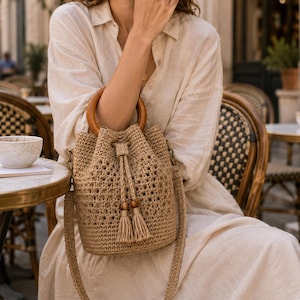 May include: A beige crochet bucket bag featuring a circular wooden handle, a drawstring closure with two tassels, and a long matching shoulder strap. The bag has an open-weave pattern and is constructed from a textured, natural-colored fiber material.