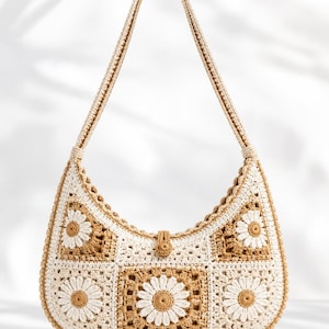 Puede incluir: Bolso de hombro de crochet crema y marrón con un estampado de flores de margarita. El bolso tiene forma curva, una correa larga y un cierre de botón. El bolso está hecho de hilo y tiene un estilo bohemio.