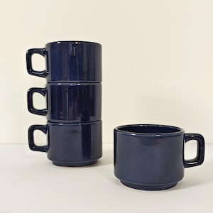Può includere: Quattro tazze in ceramica blu navy. Tre tazze sono impilate, una è separata. Ogni tazza ha un manico quadrato. Le tazze sono su una superficie bianca.
