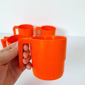 Könnte beinhalten: Mehrere stapelbare orangefarbene Kunststoffbecher mit Henkeln. Die Becher haben ein einfaches, zylindrisches Design und bestehen aus massivem, leuchtendem orangefarbenem Kunststoff. Eine Hand hält einen der Becher.