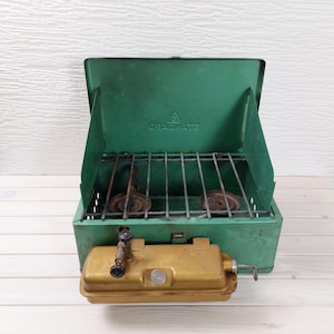 Puede incluir: Estufa de camping verde y dorada vintage con una parrilla de metal y dos quemadores. La estufa tiene una tapa con bisagras y un depósito de combustible dorado. La estufa está diseñada para cocinar al aire libre y acampar.